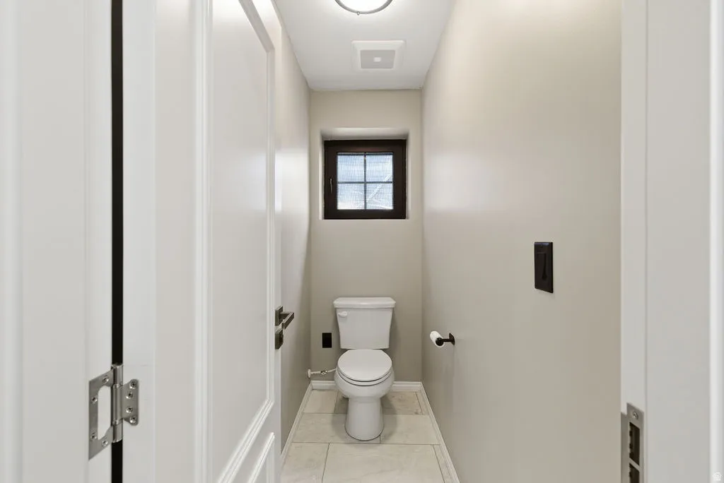 Compact powder room featuring a toilet, a small window with dark trim, light-colored wall paint, tiled flooring, and a ceiling light fixture