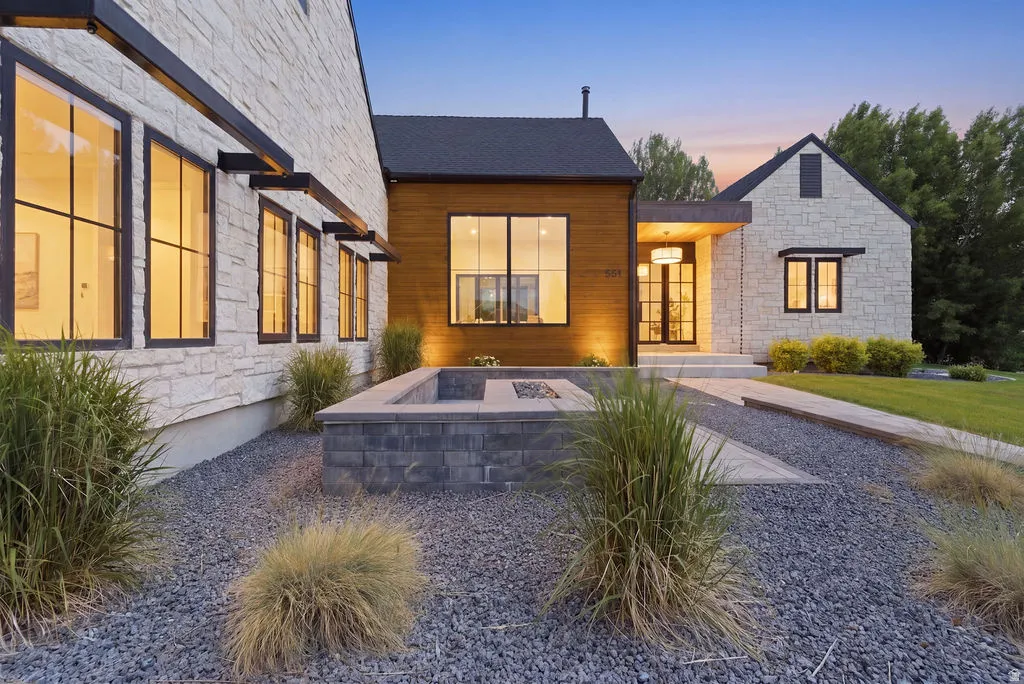 Modern exterior featuring a stone facade, wood-clad accents, a built-in fire pit, black-framed windows, and a gravel landscape