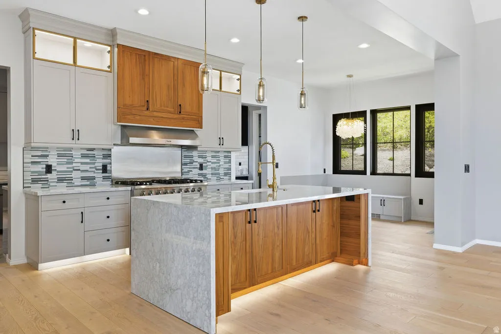 Gourmet kitchen featuring a large waterfall island with integrated wood-finish cabinetry, stainless steel range with hood, custom two-tone cabinetry with illuminated upper units, and light wood-finish flooring