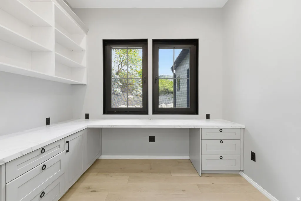 Dedicated office space featuring two windows with black frames, built-in shelving, light wood-finish flooring, and a wrap-around desk with white countertops and light gray cabinetry