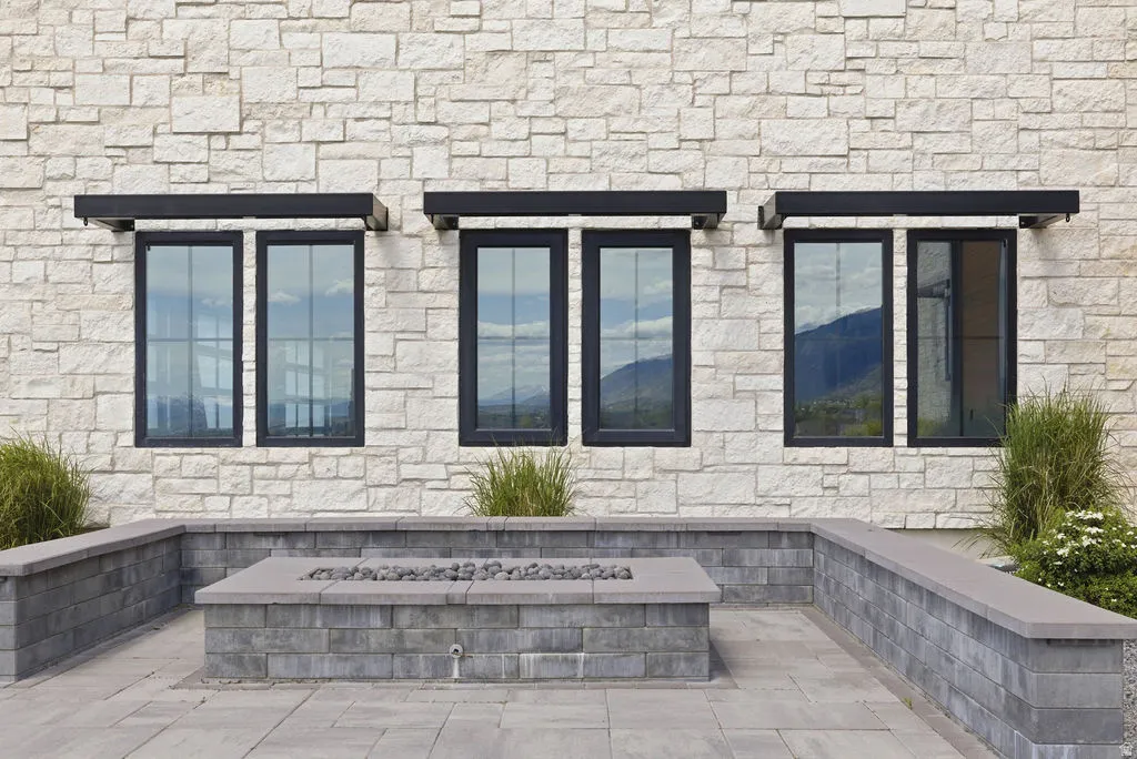 Stone exterior with dark-framed windows under black awnings, complemented by a paver patio featuring a built-in fire pit and retaining wall seating