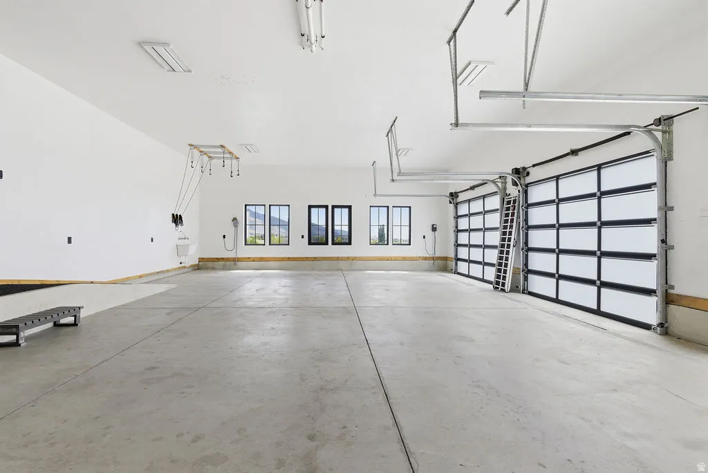 Spacious garage with concrete flooring, two contemporary overhead doors, multiple windows with dark framing, bright overhead lighting, and built-in utility connections