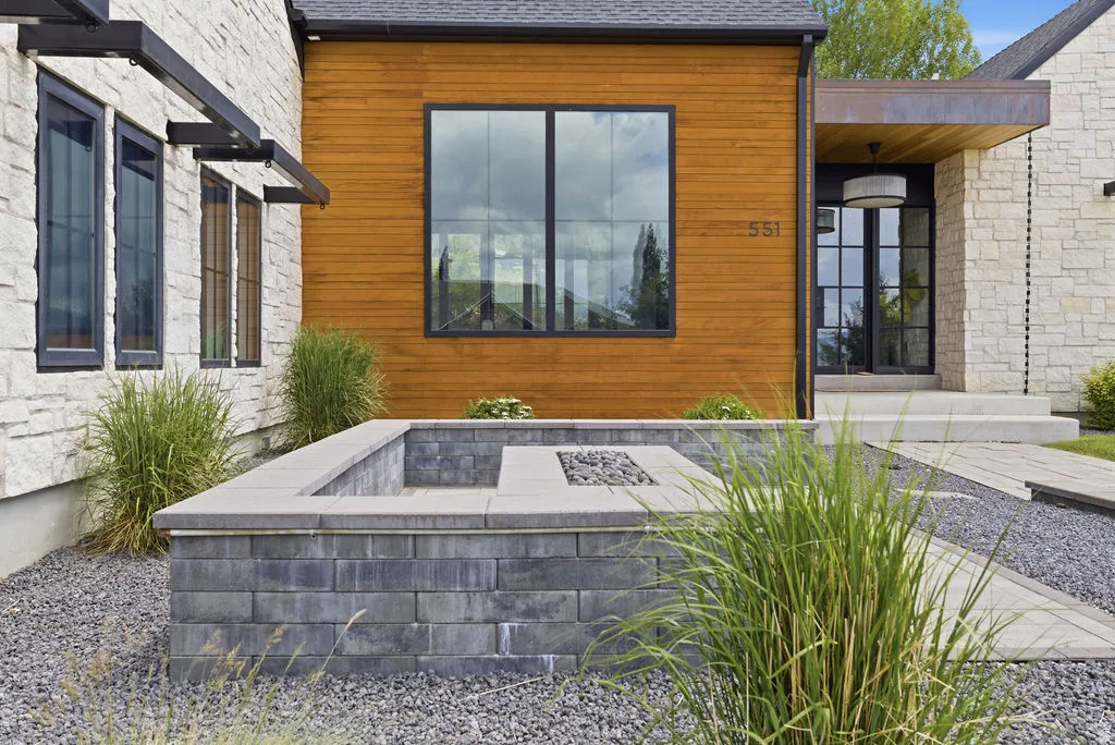 Stone and wood-clad exterior with a contemporary fire pit, dark-framed windows, and a paved walkway leading to the entrance