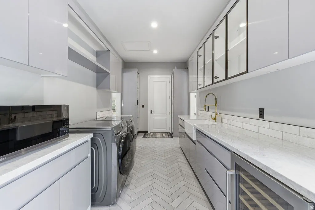 Galley-style utility room featuring light gray cabinetry, white countertops, herringbone-patterned floor tile, a built-in wine cooler, and a farmhouse-style sink with a gold-tone faucet