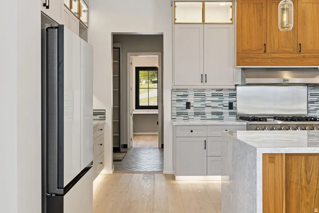 Gourmet kitchen featuring a professional-grade range with a stainless steel hood, light wood-finish flooring, a contemporary mosaic tile backsplash, custom cabinetry with integrated lighting, and a waterfall island with a light-toned countertop