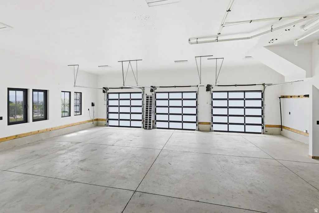 Spacious multi-car garage featuring three contemporary glass-paneled doors, concrete flooring, multiple windows with black frames, and exposed ceiling ductwork