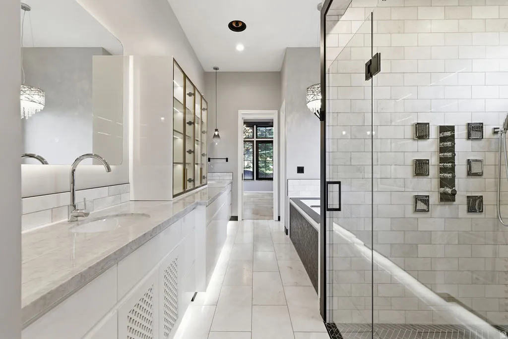 Spacious bathroom featuring dual vanity with undermount sinks and chrome fixtures, white tile flooring, a glass-enclosed shower with subway tile surround, and built-in display cabinetry