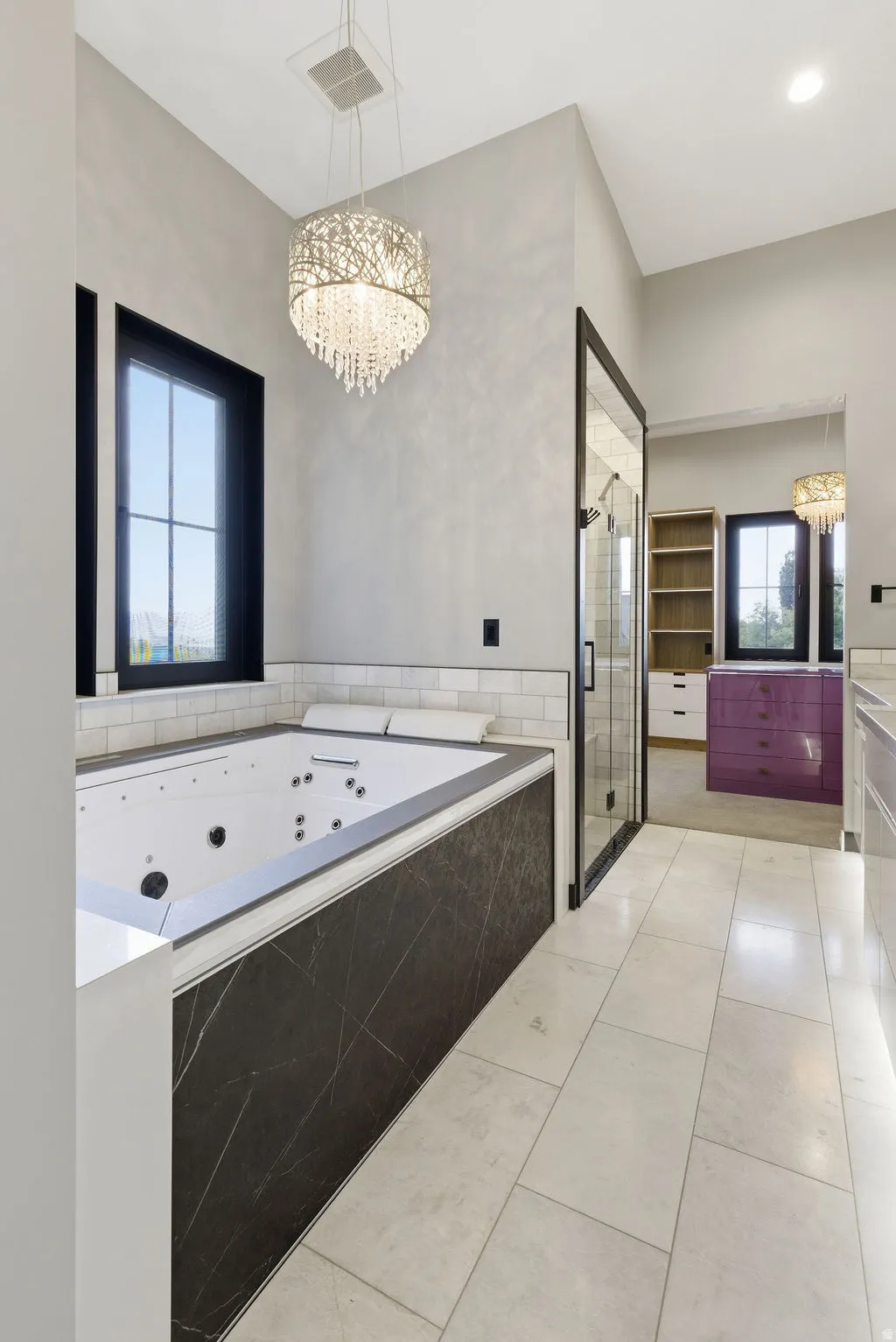 Soaking tub with a dark stone-look surround, white ceramic tile, and a decorative crystal chandelier
