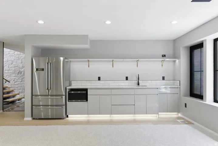 Modern wet bar featuring a stainless steel French-door refrigerator, integrated microwave, undermount sink with a matte black faucet, and light gray cabinetry with under-cabinet lighting