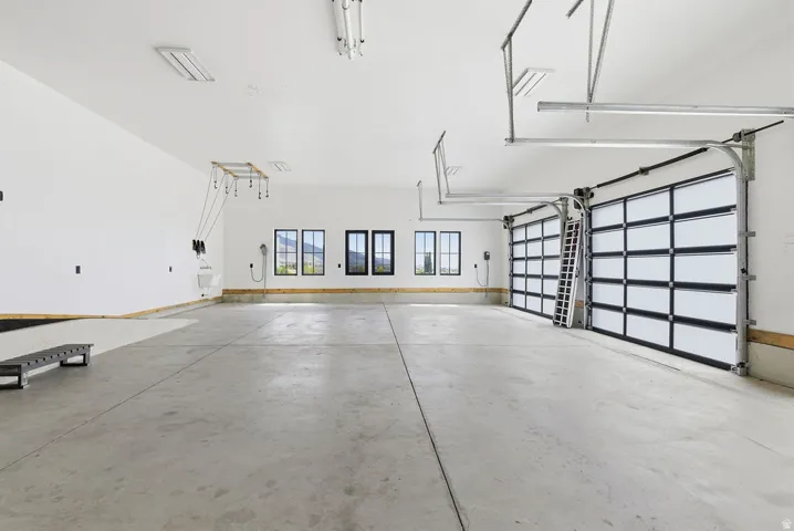 Spacious garage with concrete flooring, two contemporary overhead doors, multiple windows with dark framing, bright overhead lighting, and built-in utility connections
