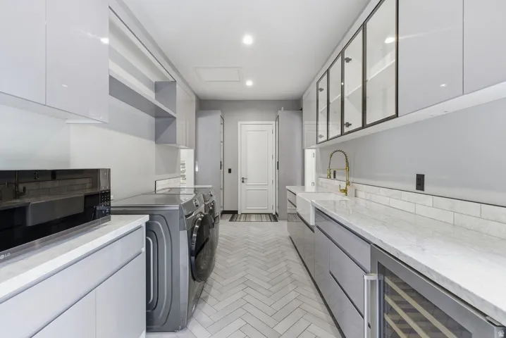 Galley-style utility room featuring light gray cabinetry, white countertops, herringbone-patterned floor tile, a built-in wine cooler, and a farmhouse-style sink with a gold-tone faucet