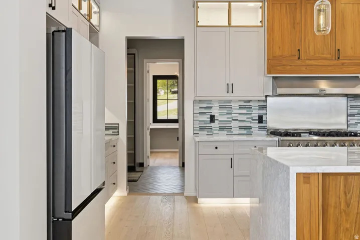 Gourmet kitchen featuring a professional-grade range with a stainless steel hood, light wood-finish flooring, a contemporary mosaic tile backsplash, custom cabinetry with integrated lighting, and a waterfall island with a light-toned countertop