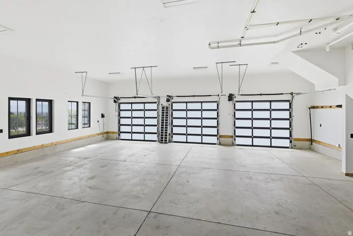 Spacious multi-car garage featuring three contemporary glass-paneled doors, concrete flooring, multiple windows with black frames, and exposed ceiling ductwork