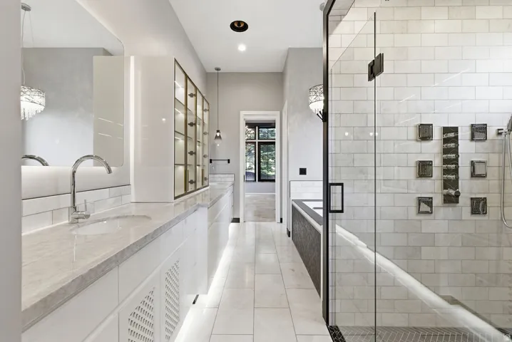 Spacious bathroom featuring dual vanity with undermount sinks and chrome fixtures, white tile flooring, a glass-enclosed shower with subway tile surround, and built-in display cabinetry