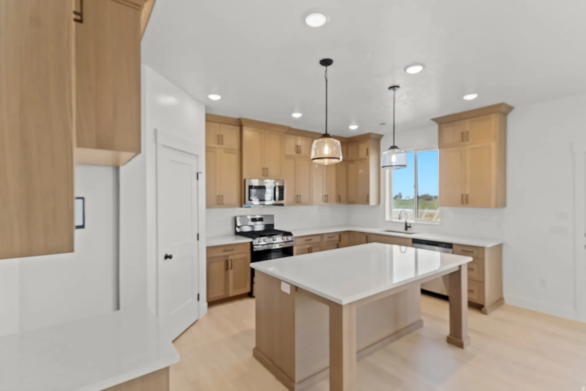 Spacious kitchen featuring light wood-finish cabinetry, a large center island with a light-colored countertop, stainless steel appliances, and pendant lighting