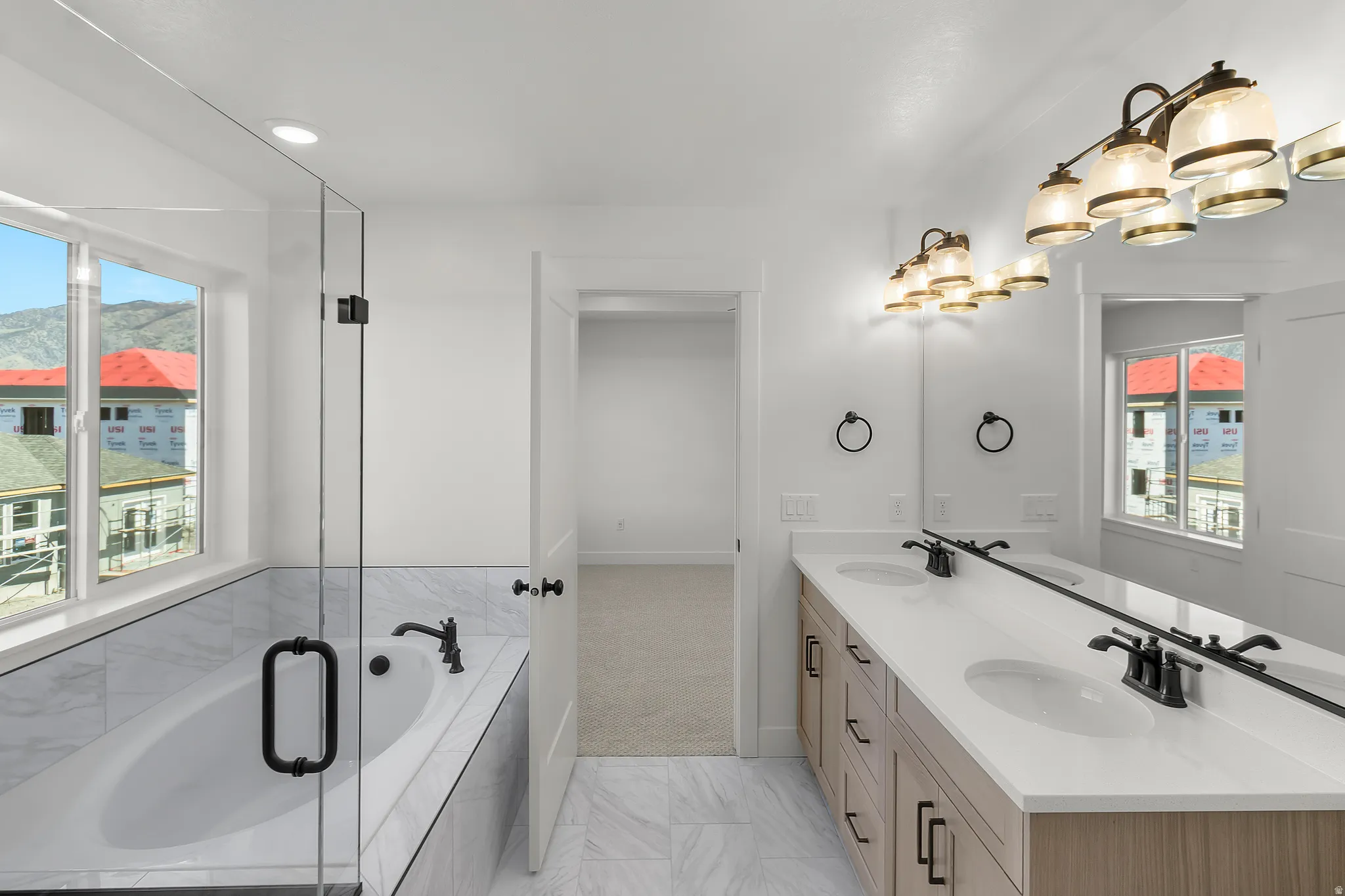 Bathroom featuring a built-in soaking tub with surrounding tile, a frameless glass shower enclosure, and a dual-sink vanity with light wood-finish cabinetry, white countertops, and matte black hardware