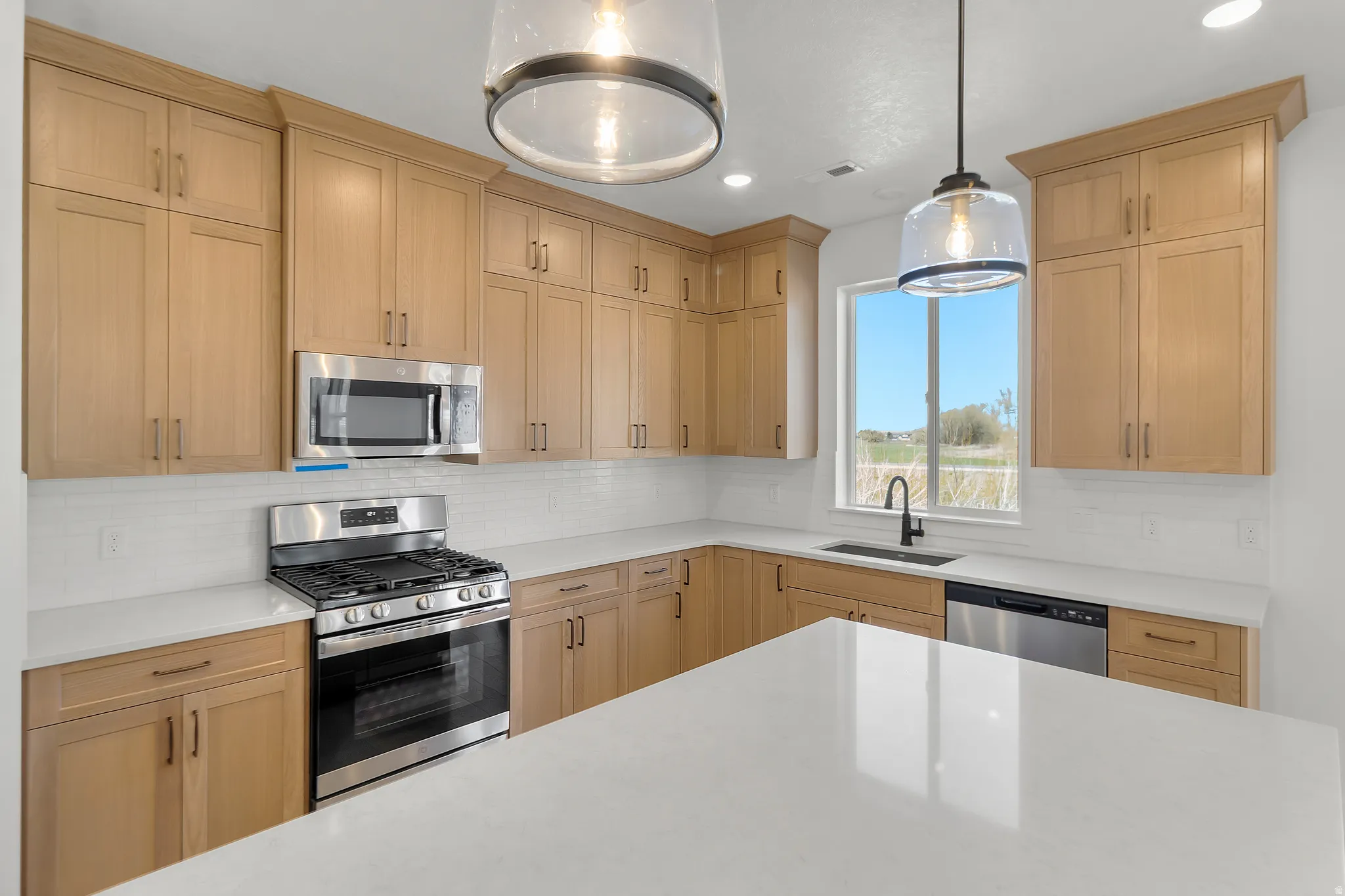 Modern kitchen featuring light wood-finish cabinetry, stainless steel appliances, a white tile backsplash, a large center island with a white countertop, and contemporary pendant lighting