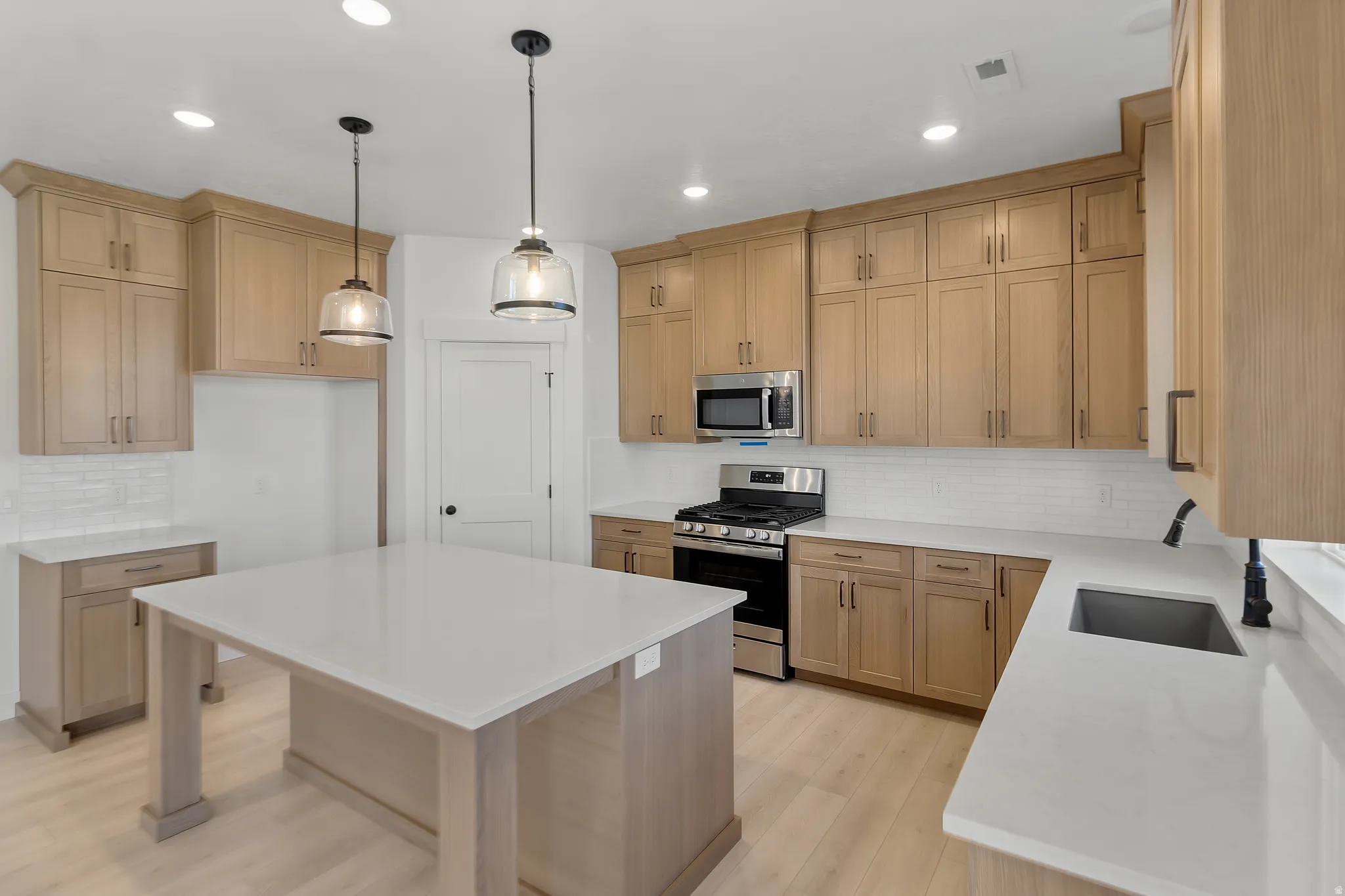 Kitchen featuring a central island with a light-toned countertop, wood-finish flooring, light wood cabinetry, stainless steel appliances, and subway tile backsplash