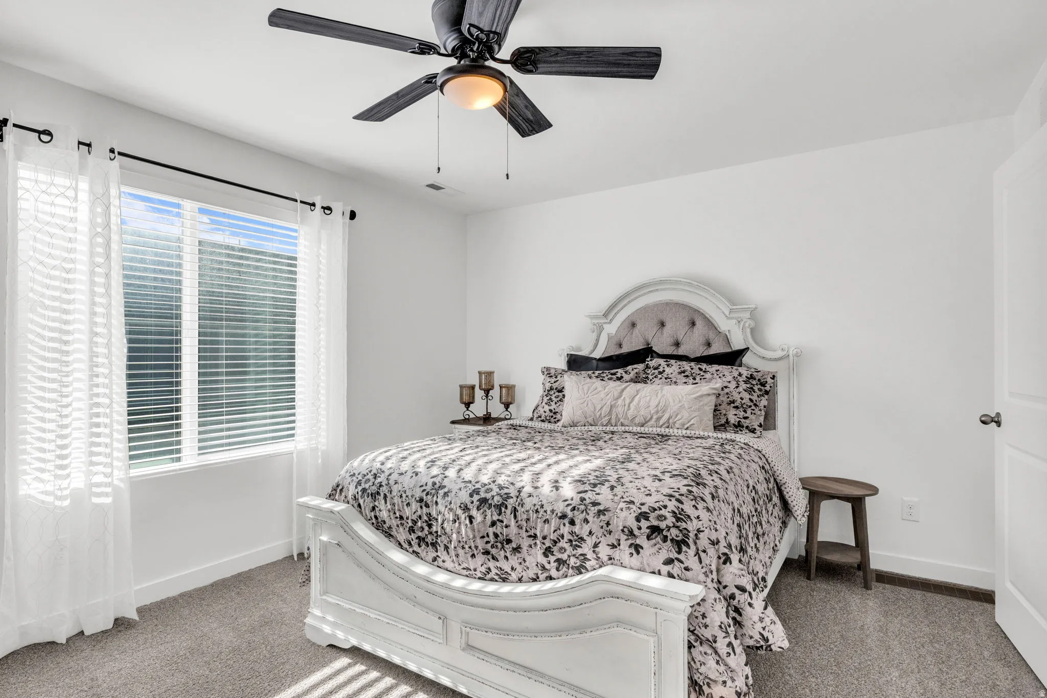 Bedroom featuring light gray carpeting, crisp white walls, a large window with blinds, and a black ceiling fan with an integrated light fixture