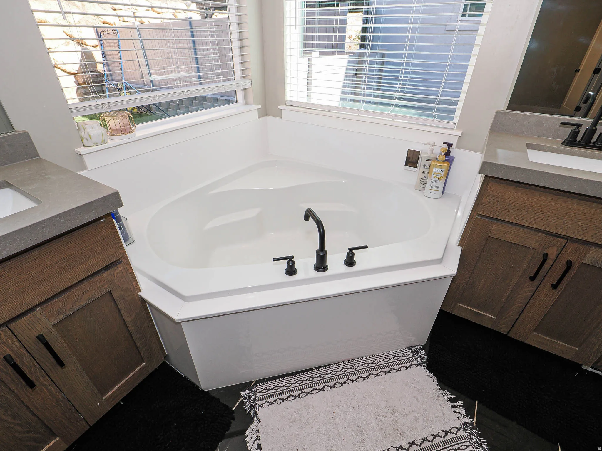 Corner soaking tub with a matte black faucet, flanked by dual wood-finish vanities with undermount sinks and stone countertops