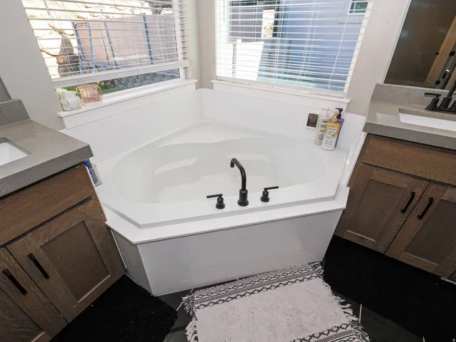 Corner soaking tub with a matte black faucet, flanked by dual wood-finish vanities with undermount sinks and stone countertops