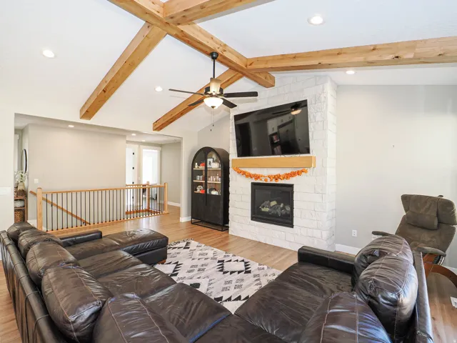 Vaulted ceiling with exposed wood beams, white brick fireplace with wood mantel, recessed lighting, ceiling fan, and wood-finish flooring