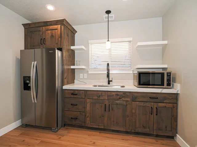 Compact kitchen area featuring wood-finish cabinetry with black hardware, stainless steel appliances, a single basin sink with a matte black faucet, and white countertops