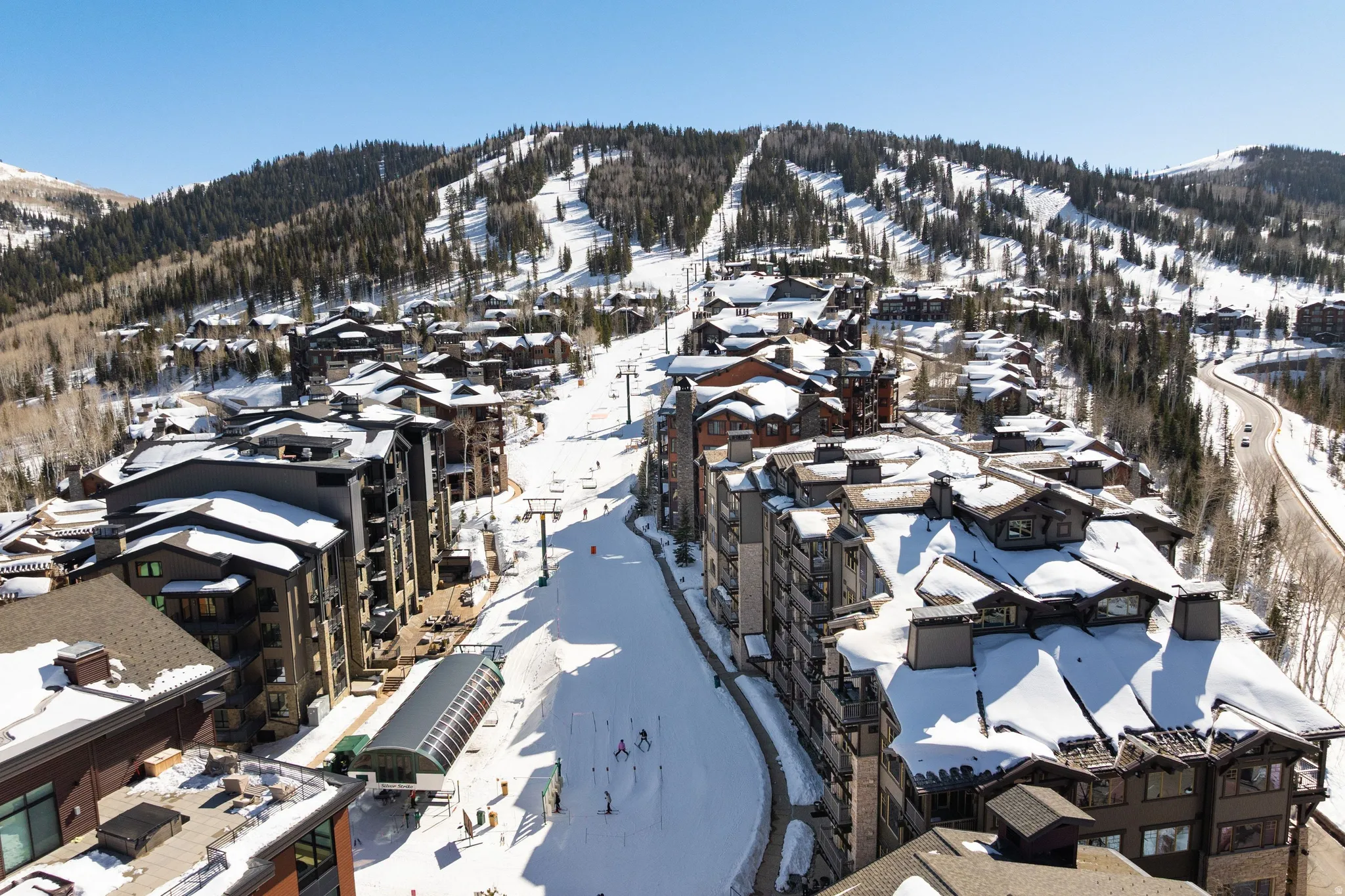 Mountain resort community featuring multi-story structures, ski slopes, mature evergreen trees, and a winding roadway