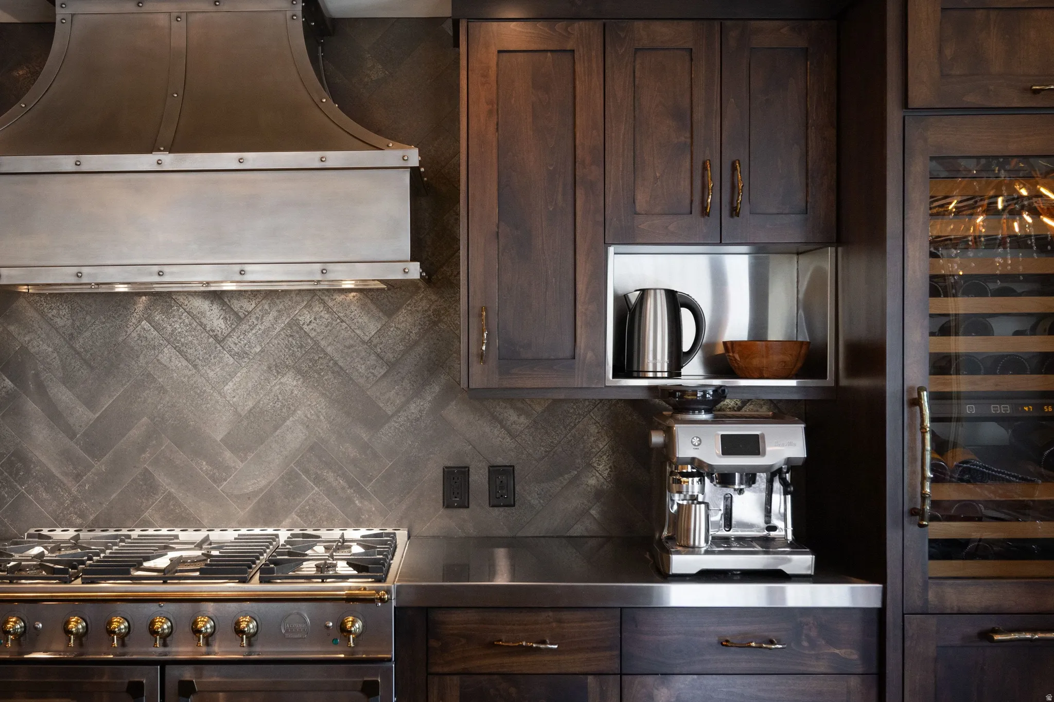Professional-grade range with brass accents, stainless steel range hood, herringbone pattern backsplash, dark wood cabinetry, and a built-in wine refrigerator