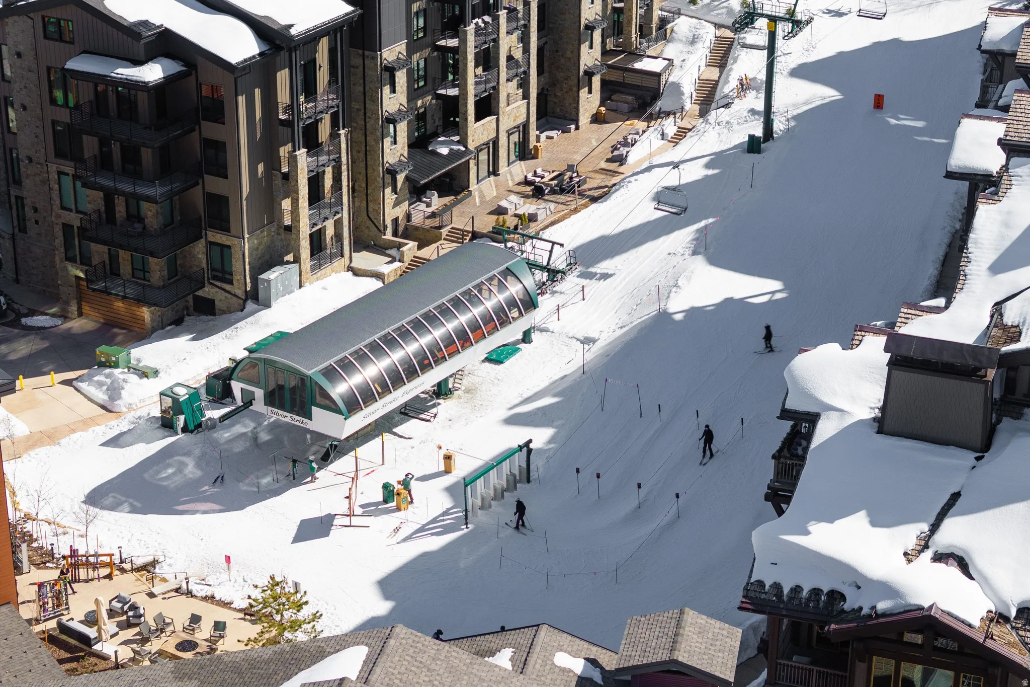 Ski resort setting featuring a ski lift base station, adjacent snow-covered slopes, and multi-story buildings with balconies