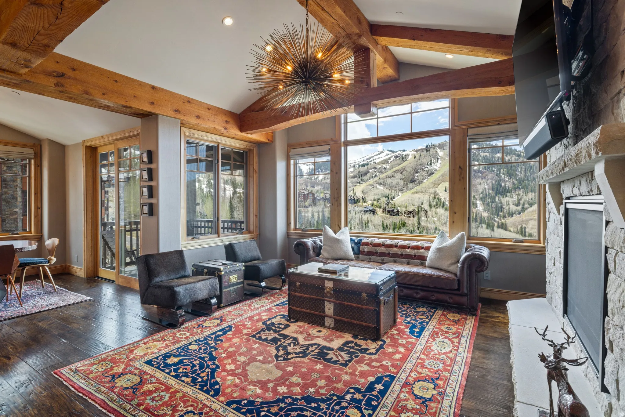 Open living area featuring expansive windows, exposed wood beams, a stone fireplace, wood-finish flooring, and a contemporary chandelier