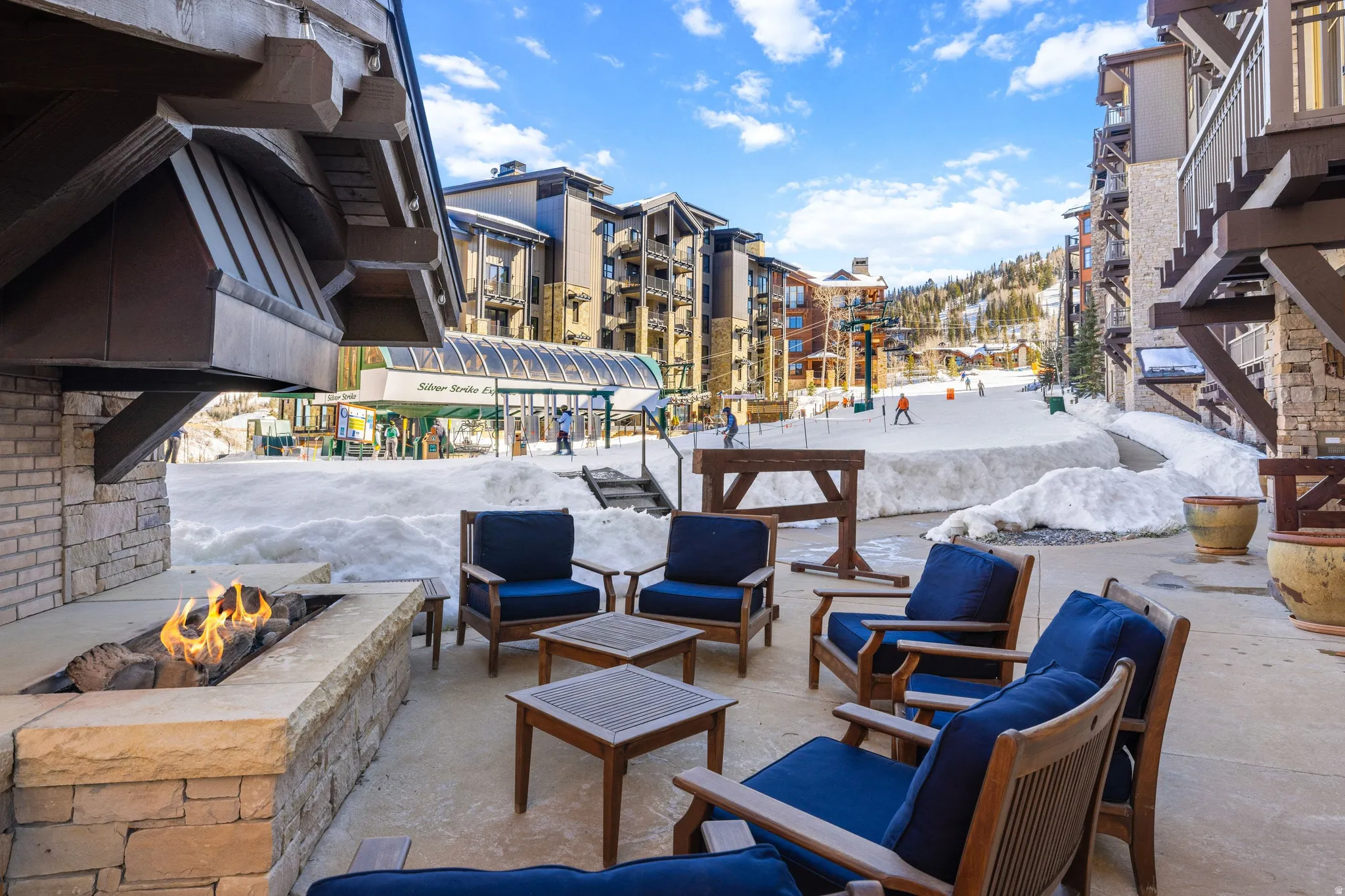 Paved patio featuring a built-in stone fire pit, wood-slat chairs with blue cushions, and a ski resort backdrop with multiple buildings
