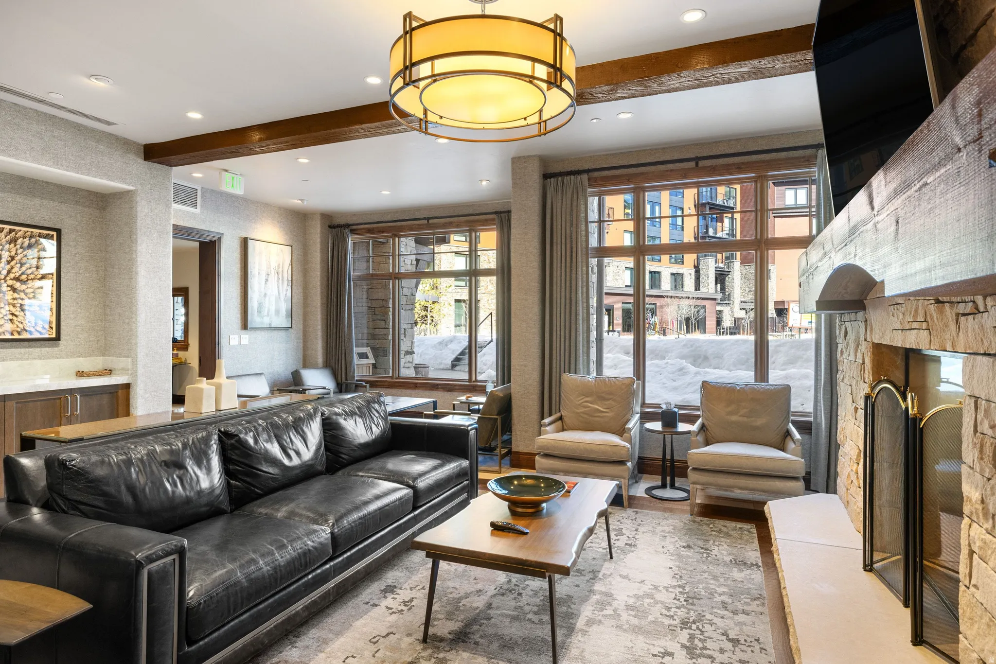 Spacious living area featuring a stone-clad fireplace with hearth, large windows, a decorative ceiling light fixture, and exposed wood beam accents