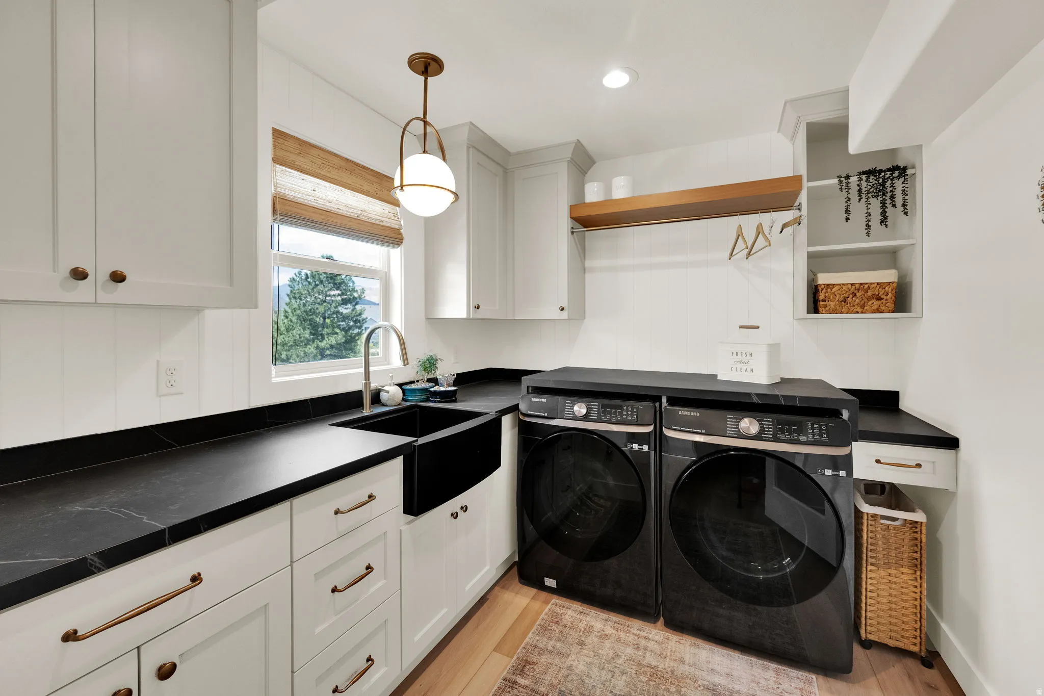 Laundry room featuring white cabinetry, black countertops, a black farmhouse sink, wood-finish flooring, and a window with a woven shade