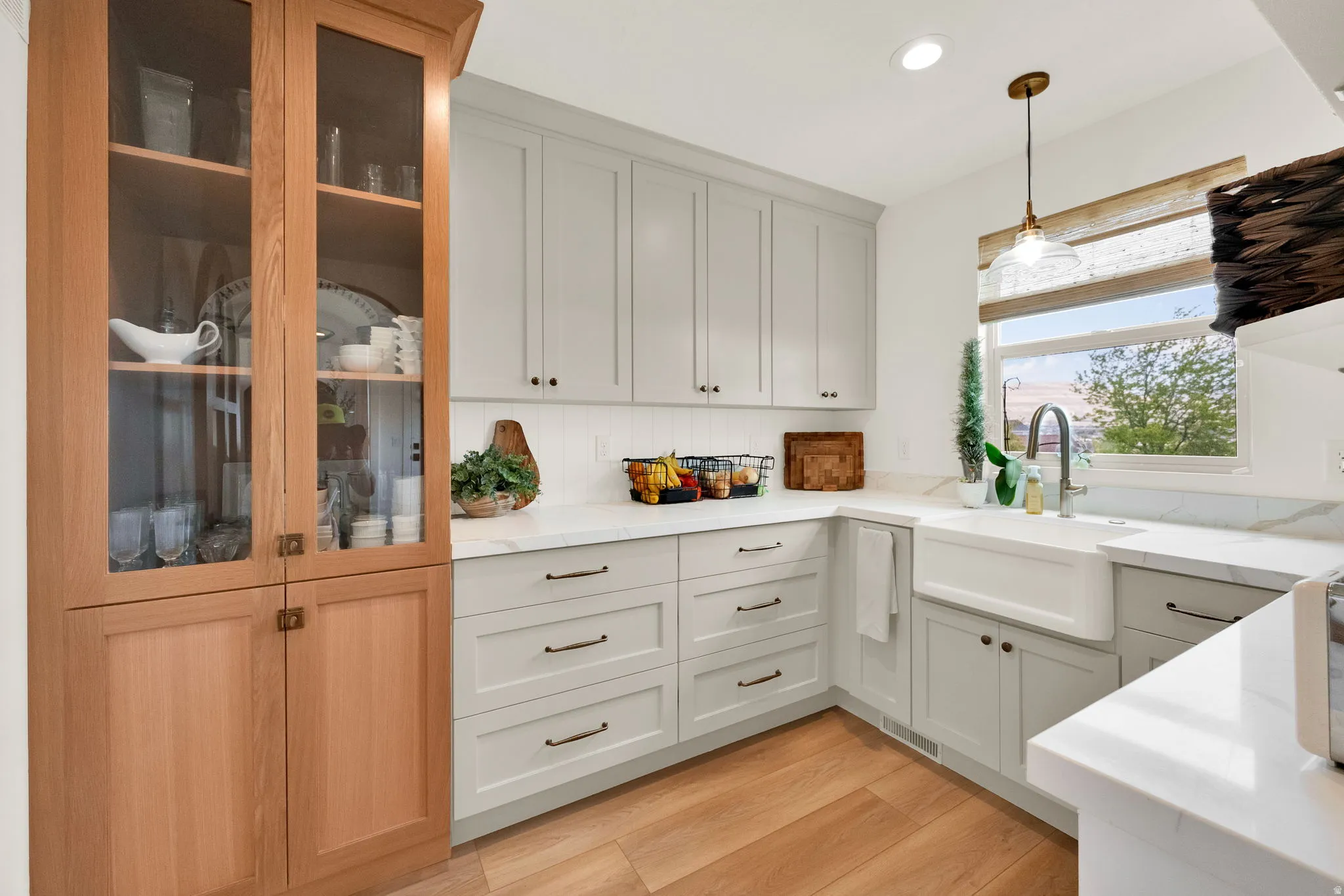 Modern kitchen featuring light wood-finish flooring, white shaker-style cabinetry, a farmhouse sink with a gooseneck faucet, and light countertops with a matching backsplash
