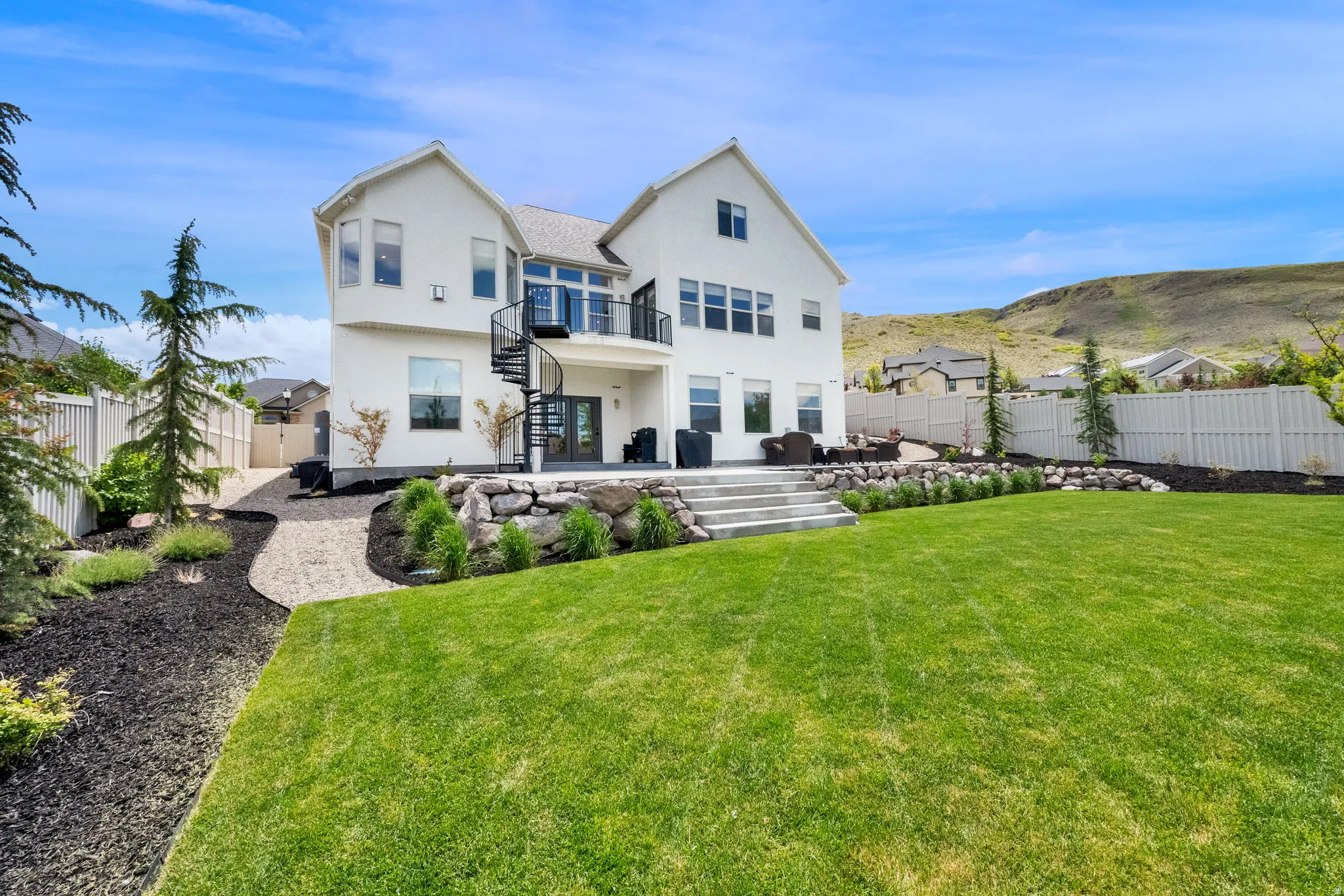 Expansive rear exterior featuring a white facade, a multi-level patio with steps, a spiral staircase leading to an upper balcony, and a landscaped yard with a lawn and mature plantings