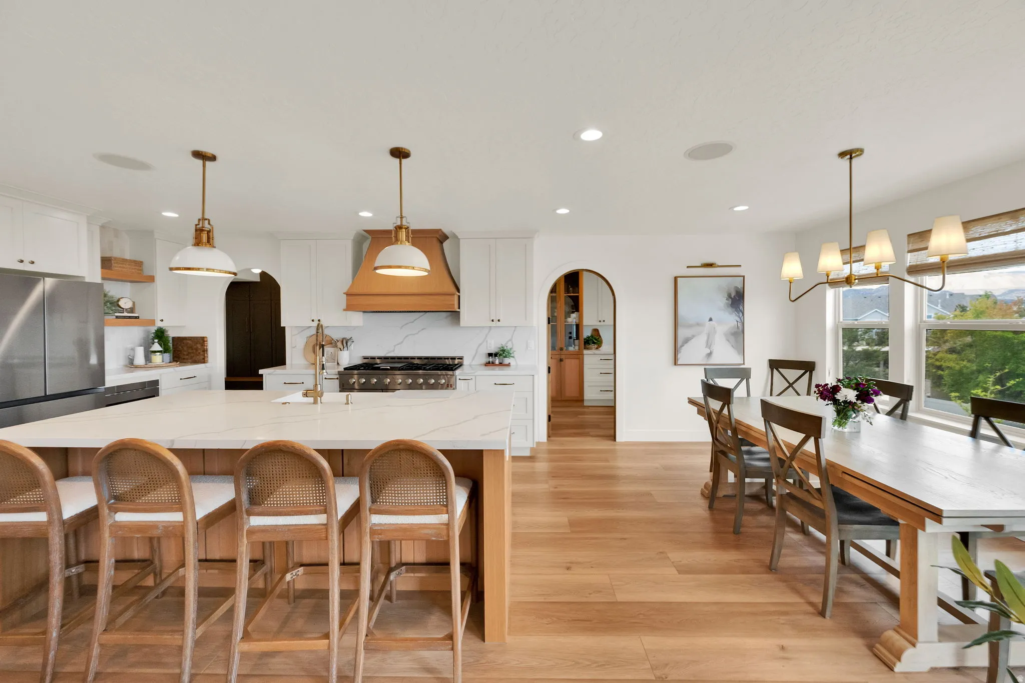 Open-concept kitchen and dining area featuring a large center island with seating, stainless steel appliances, a custom wood range hood, and light wood-finish flooring throughout