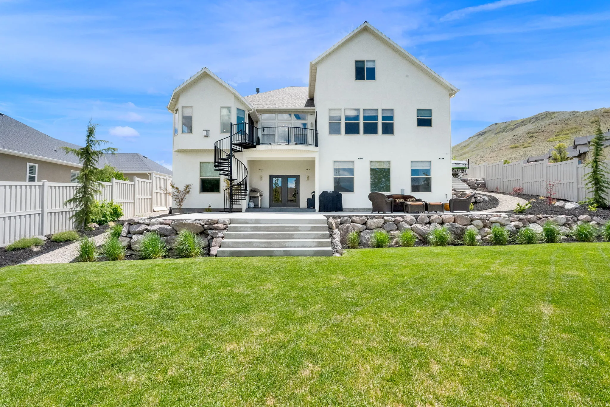 Spacious backyard featuring a manicured lawn, a large concrete patio with steps, and a rock retaining wall