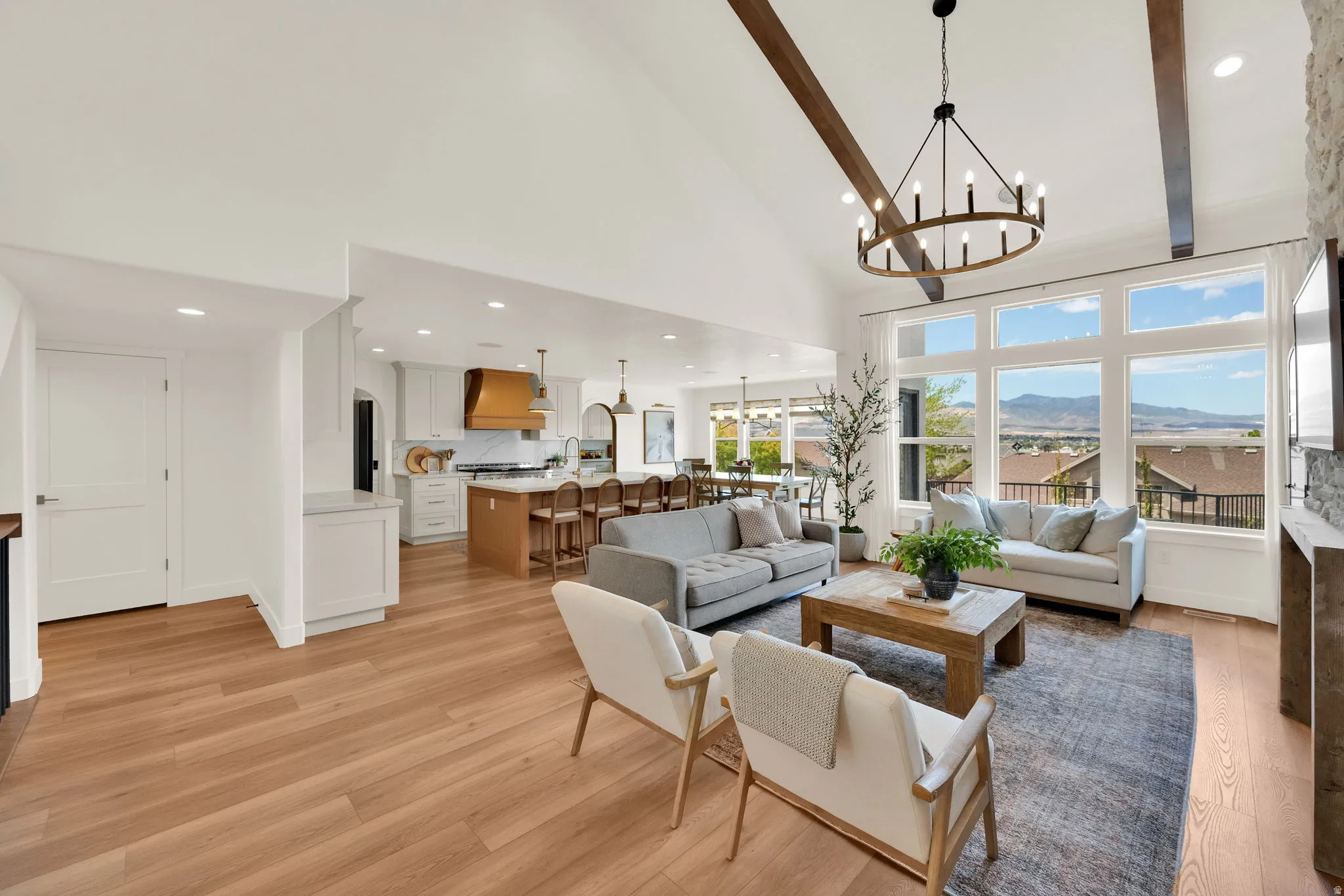 Open-concept living area featuring vaulted ceilings with exposed beams, wood-finish flooring, a contemporary chandelier, and large windows