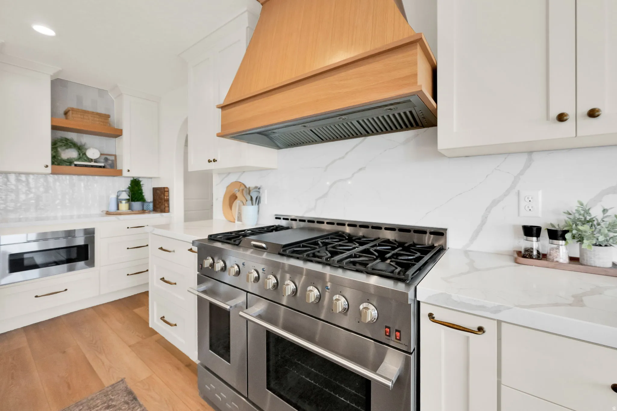 Gourmet kitchen featuring a stainless steel gas range, a natural wood-finish range hood, white cabinetry with brass hardware, and white countertops with matching slab backsplash