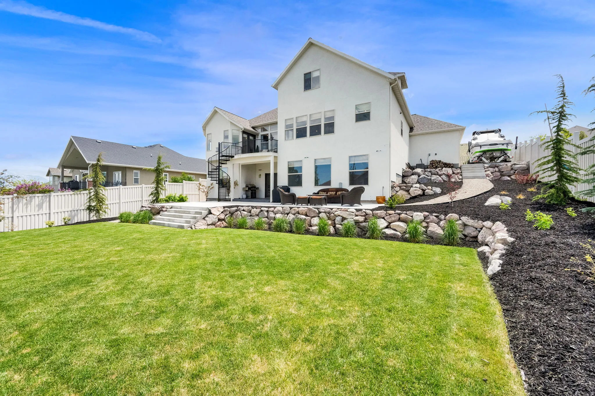 Expansive green lawn with a white stucco exterior residence featuring multiple windows, a spiral staircase, a lower-level patio, and a terraced rock landscape with dark mulch