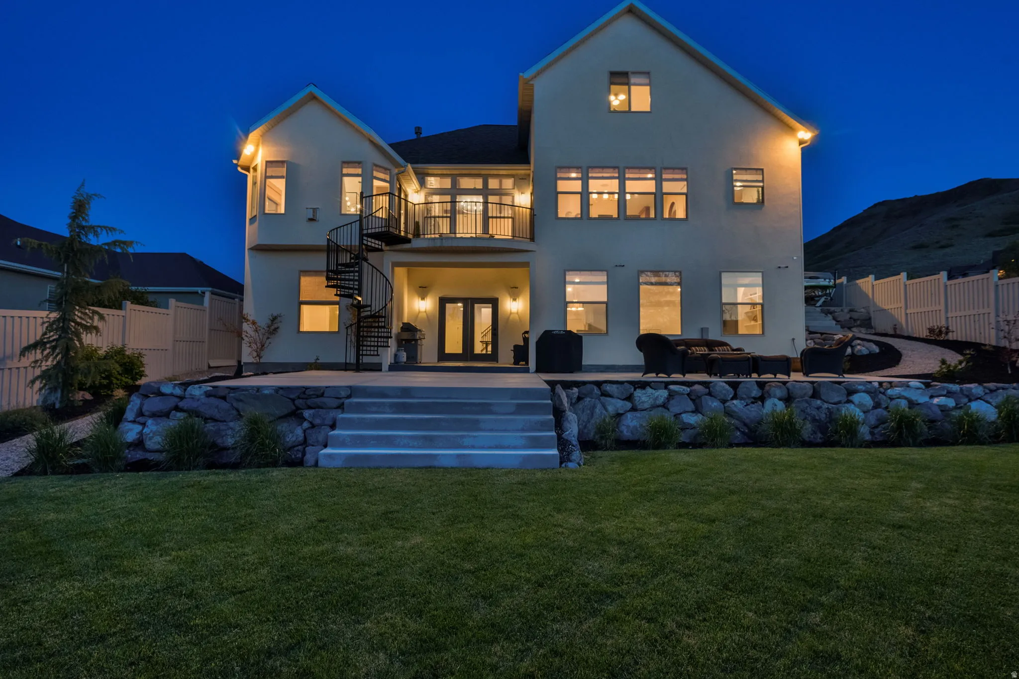 Rear exterior featuring multiple windows, a second-story balcony with a spiral staircase, a ground-level patio, and a stone retaining wall