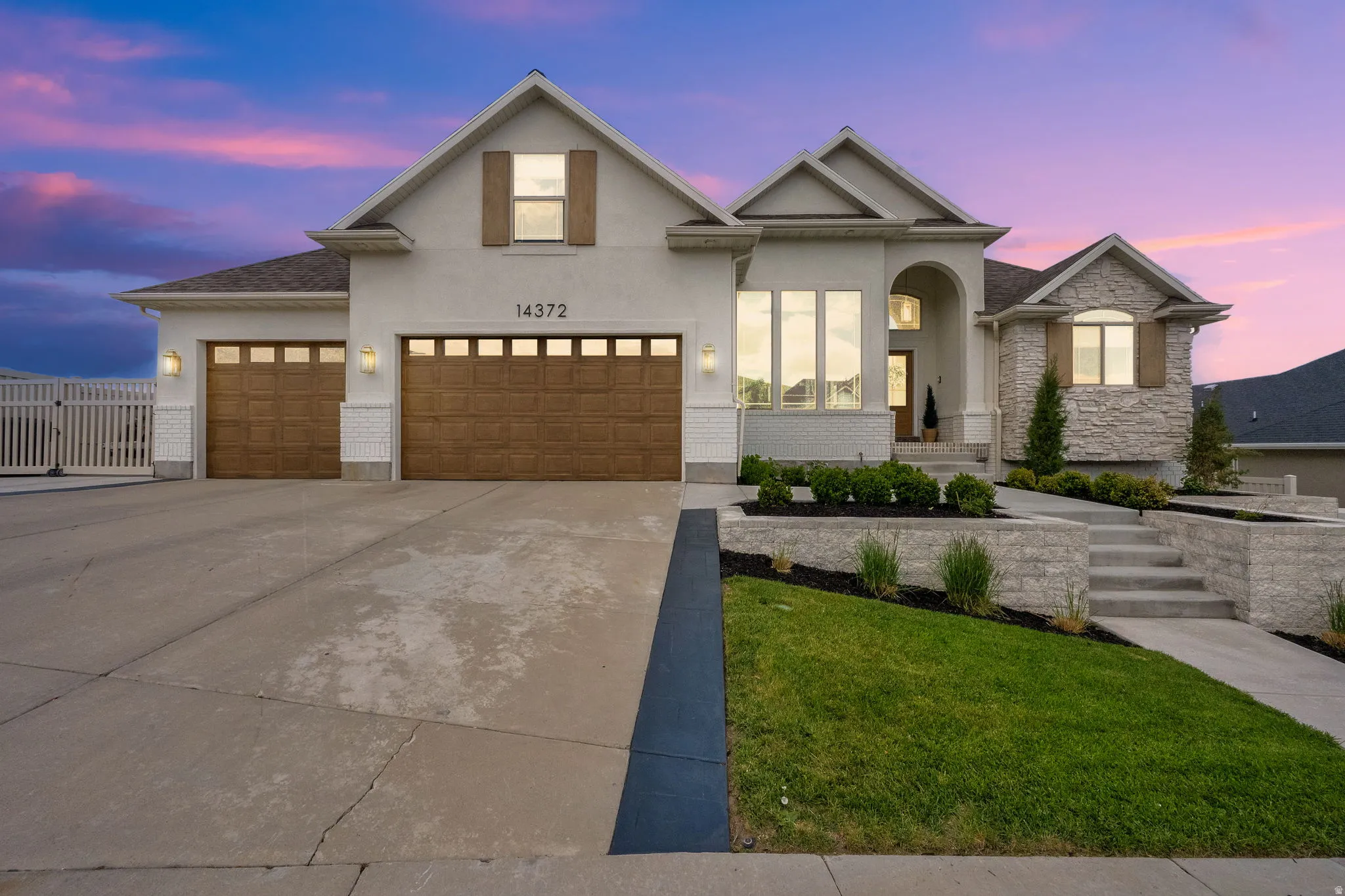 Modern stucco and stone exterior featuring two wooden-paneled garage doors, a concrete driveway, and tiered landscaping with a stone retaining wall