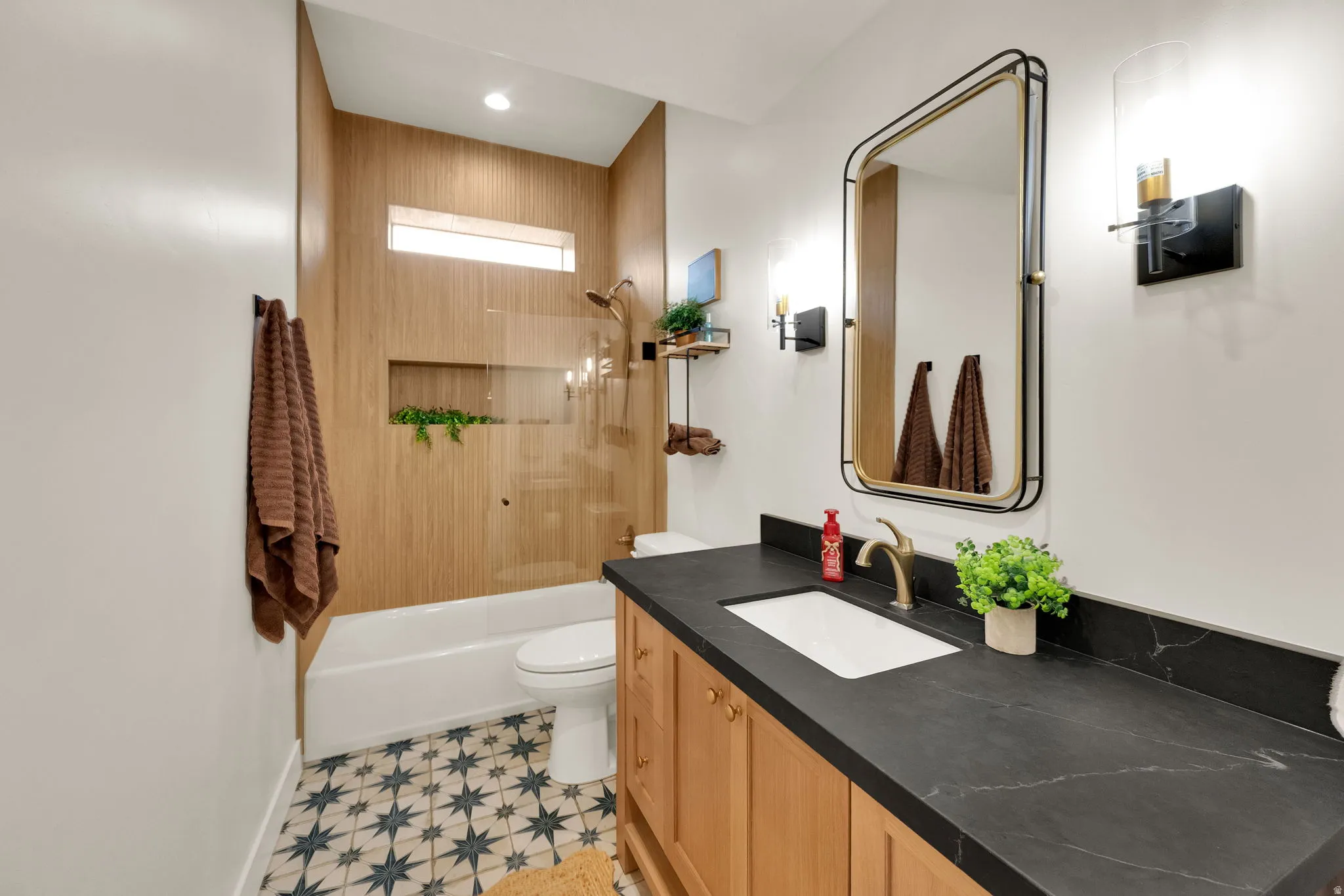 Bathroom featuring a single vanity with a dark countertop and undermount sink, patterned floor tile, a framed mirror, wall-mounted sconces, and a shower-tub combination with wood-finish wall panels and a glass enclosure