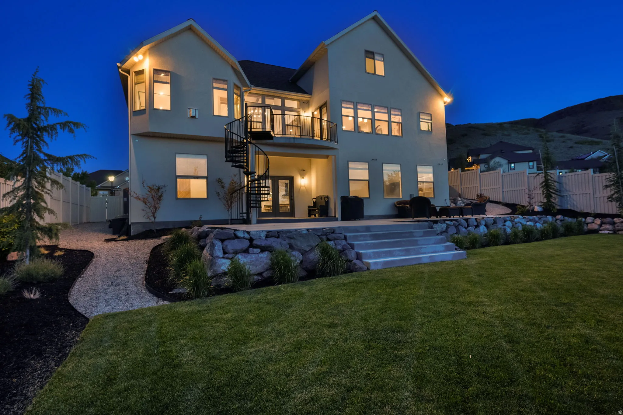 Expansive rear facade featuring a second-story balcony with a spiral staircase, a ground-level patio, a tiered retaining wall, and a manicured lawn