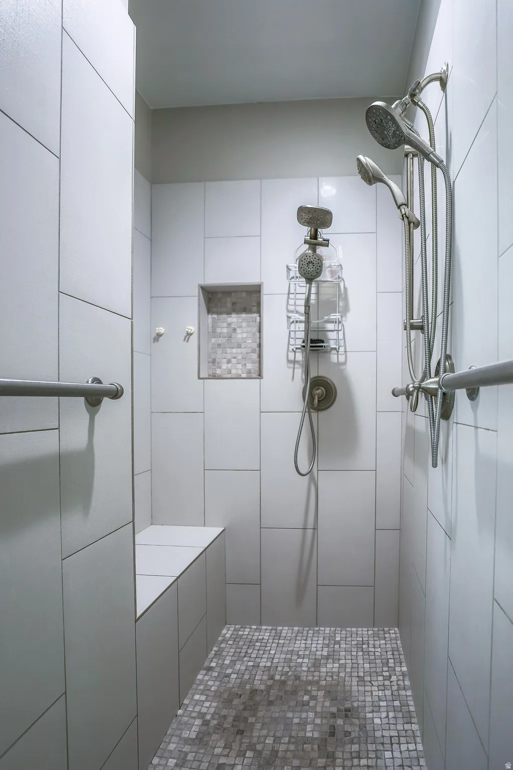 Spacious tiled shower featuring a built-in bench, recessed niche with mosaic tile, dual shower heads, and mosaic tile flooring