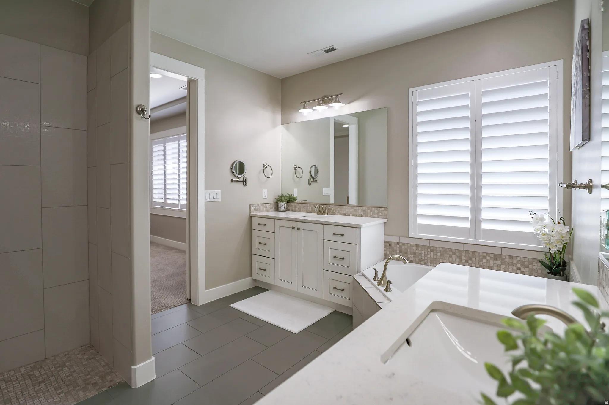 Bathroom featuring a dual vanity with white cabinetry, a built-in bathtub with mosaic tile surround, plantation shutters, and a walk-in shower with tile walls
