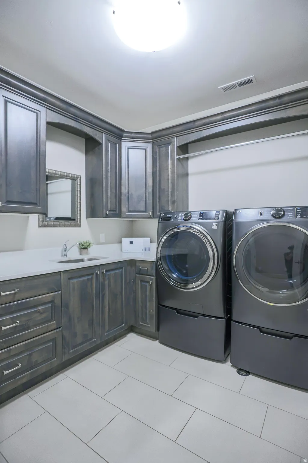 Laundry area featuring extensive dark wood cabinetry, light-toned countertops, a stainless steel sink with a gooseneck faucet, and light rectangular floor tiling