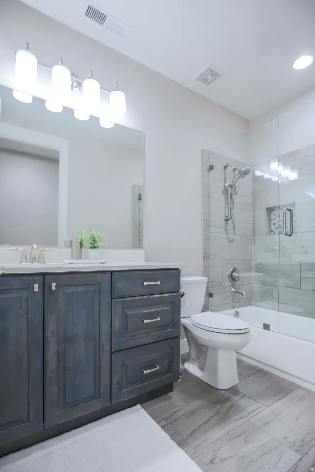 Modern bathroom featuring a wood-finish vanity with a light countertop, a built-in tub with a glass enclosure, and large format tile flooring