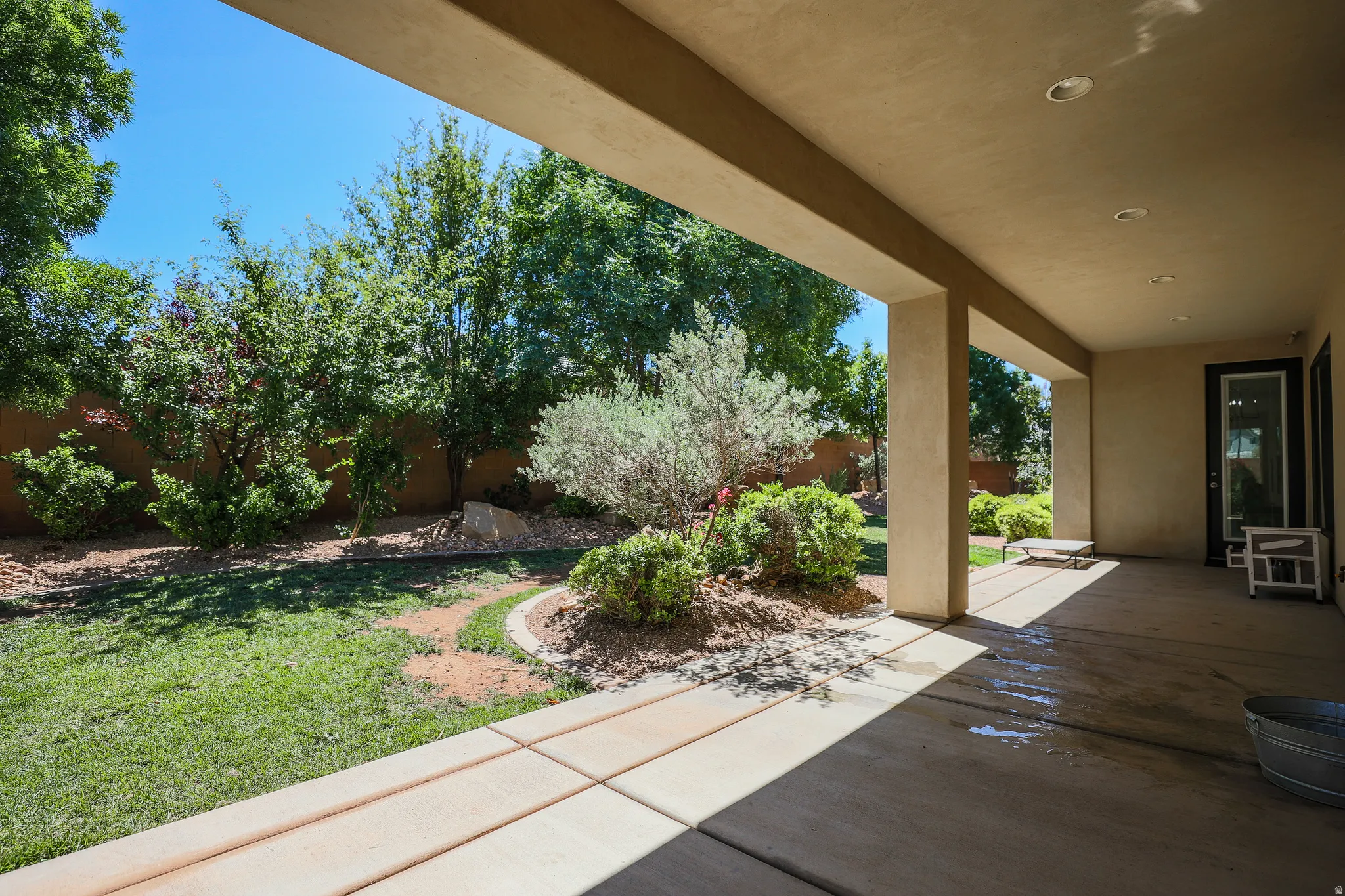 Spacious covered patio with recessed lighting overlooking a landscaped backyard featuring mature trees, shrubs, and a curved stone-edged garden bed